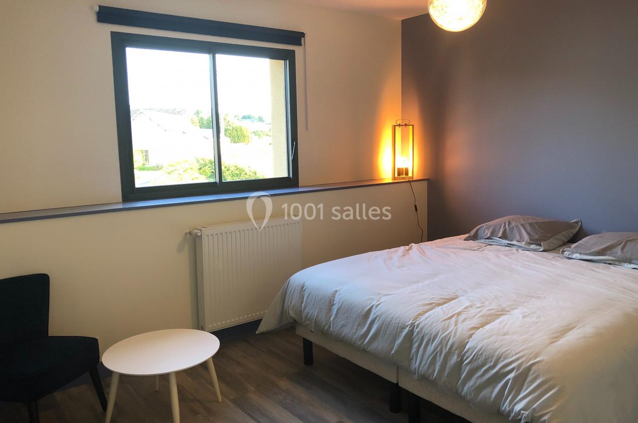 Chambre lumineuse avec un lit double, une table basse, un fauteuil, un mur gris et une fenêtre donnant sur l'extérieur.