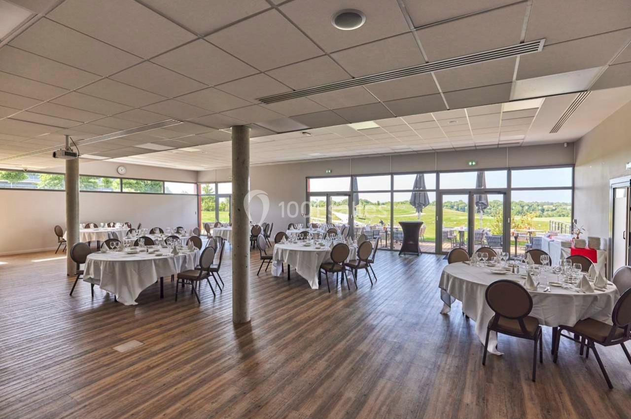 Salle de réception lumineuse avec tables rondes dressées, grandes baies vitrées donnant sur un paysage verdoyant.