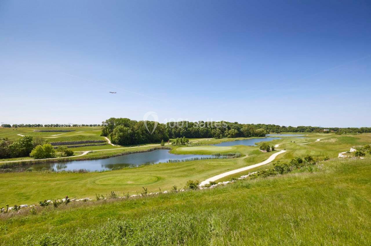 Paysage de golf avec étangs, pelouses verdoyantes, sentiers sinueux et arbres sous un ciel bleu dégagé.