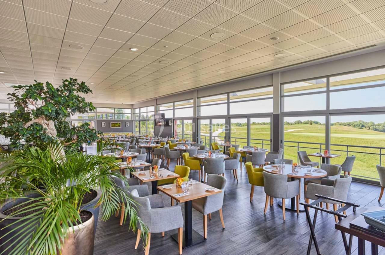 Salle de restaurant lumineuse avec de grandes baies vitrées donnant sur un paysage verdoyant, tables dressées.