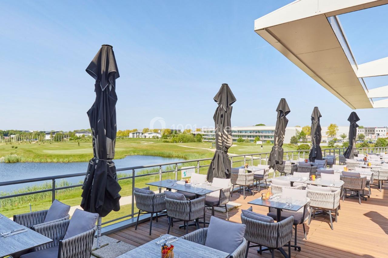 Terrasse d'un restaurant avec tables et parasols, offrant une vue sur un lac et un paysage verdoyant par temps ensoleillé.