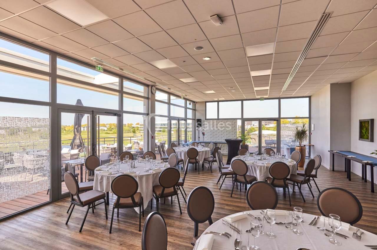 Salle lumineuse avec tables rondes dressées, grandes baies vitrées donnant sur une terrasse et vue extérieure dégagée.