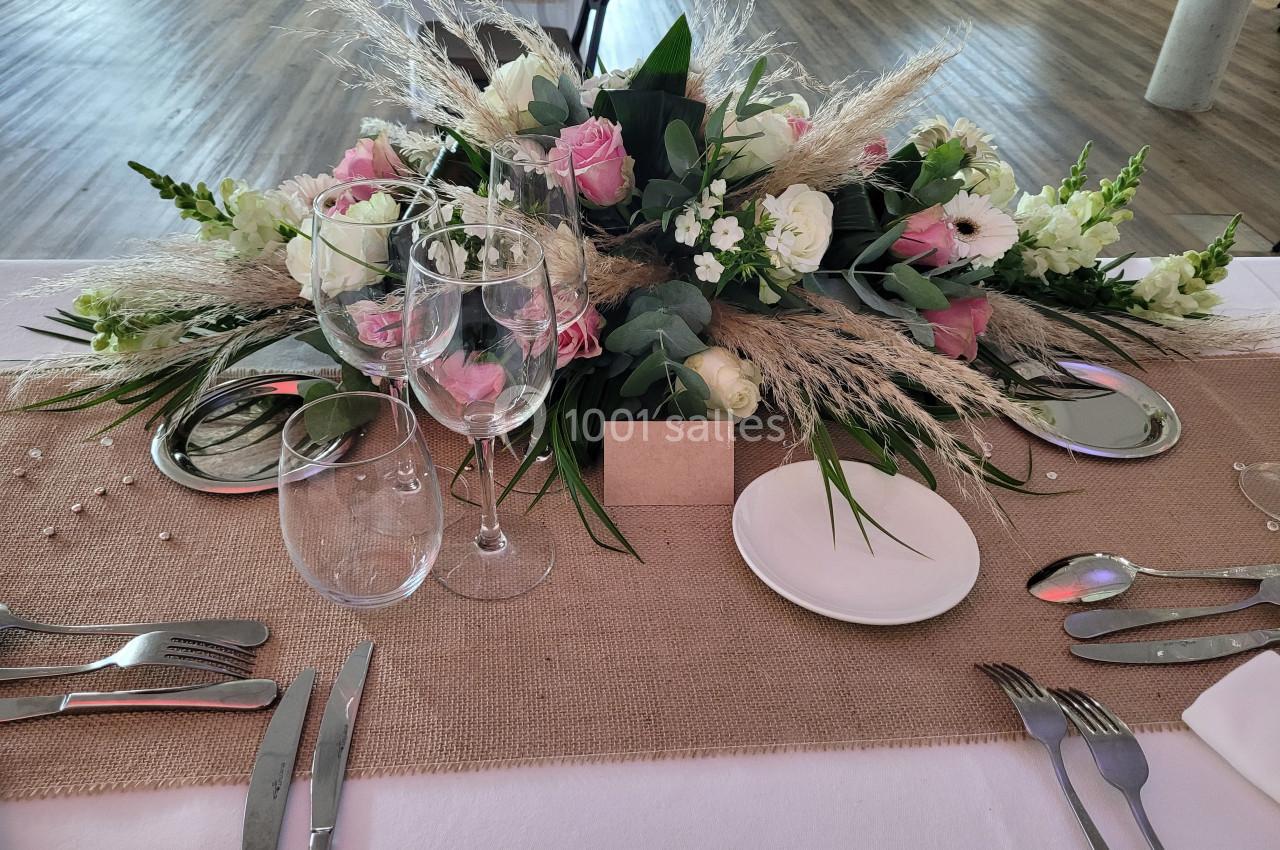 Centre de table avec fleurs roses et blanches, verres à vin, assiettes et couverts disposés sur une nappe beige.