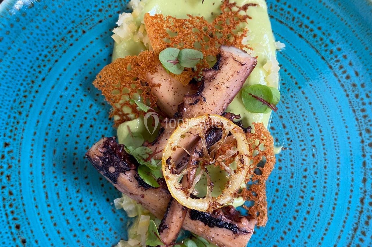 Assiette de poulpe grillé avec citron, purée verte, chips croustillantes et herbes fraîches, servie sur une assiette bleue.