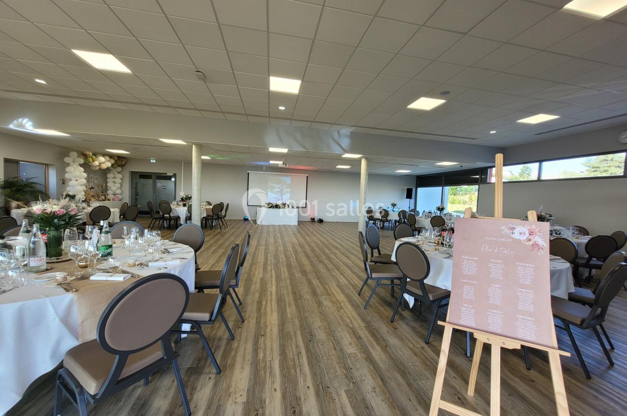 Salle de réception lumineuse avec tables dressées pour un événement et un plan de table sur un chevalet au premier plan.
