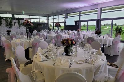 Salle de réception décorée avec des tables dressées, des chaises ornées de nœuds roses et des bouquets de fleurs.