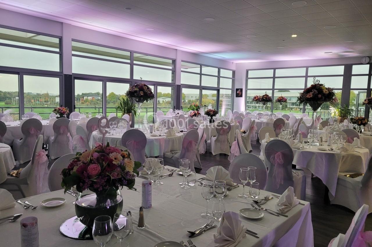 Salle de réception décorée avec des tables dressées, des chaises ornées de nœuds roses et des bouquets de fleurs.