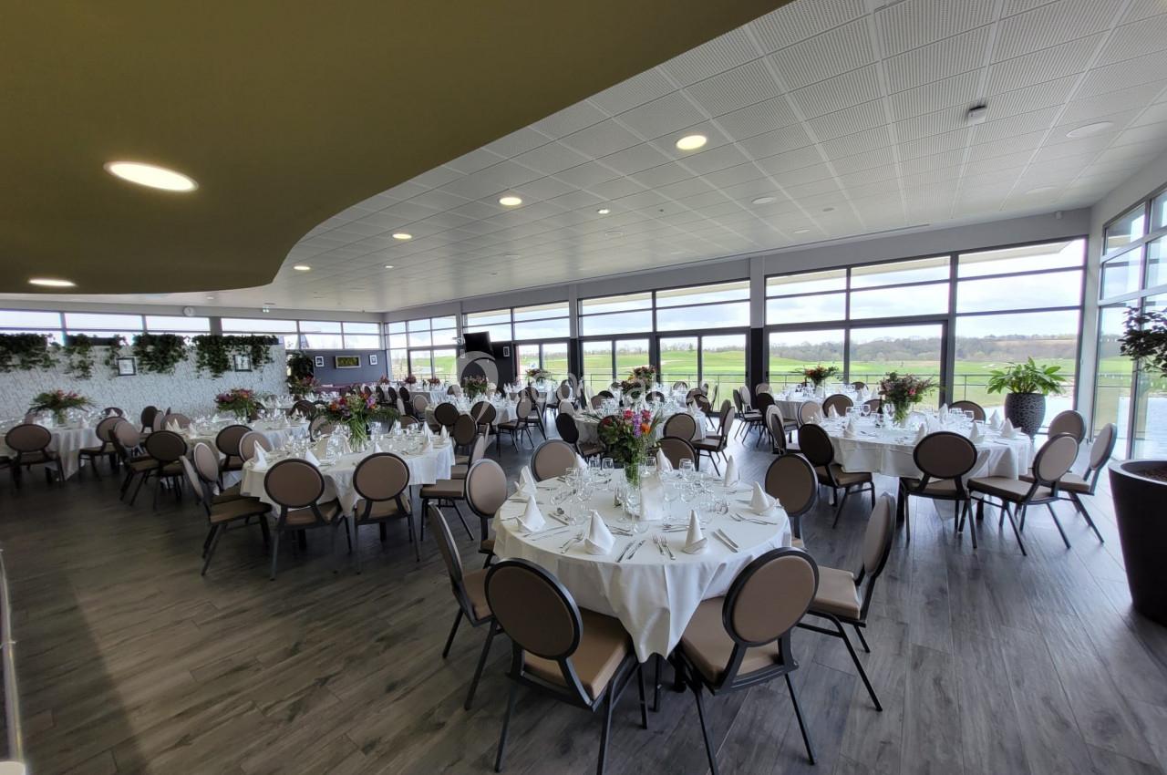Salle lumineuse avec de grandes baies vitrées, tables rondes dressées avec nappes blanches et décorations florales.