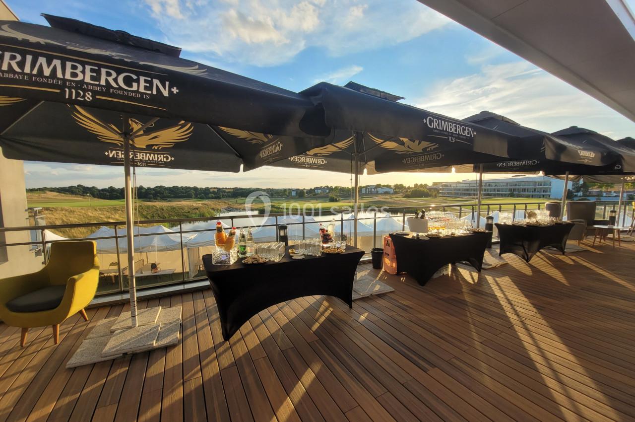 Terrasse en bois avec tables dressées sous des parasols noirs, offrant une vue sur un paysage naturel au coucher du soleil.