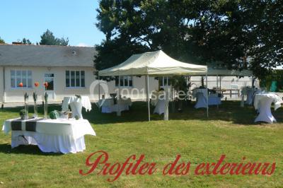 Tente blanche montée sur une pelouse, équipée de tables hautes, devant un bâtiment clair et des arbres.