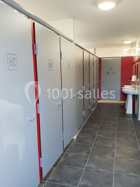 Alignement de cabines de toilettes grises avec portes fermées, lavabo et miroirs visibles au fond.