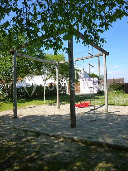 Structure de jeux en bois avec échelle et cordes, installée sur un sol sablonneux entouré d'arbres et d'herbe.