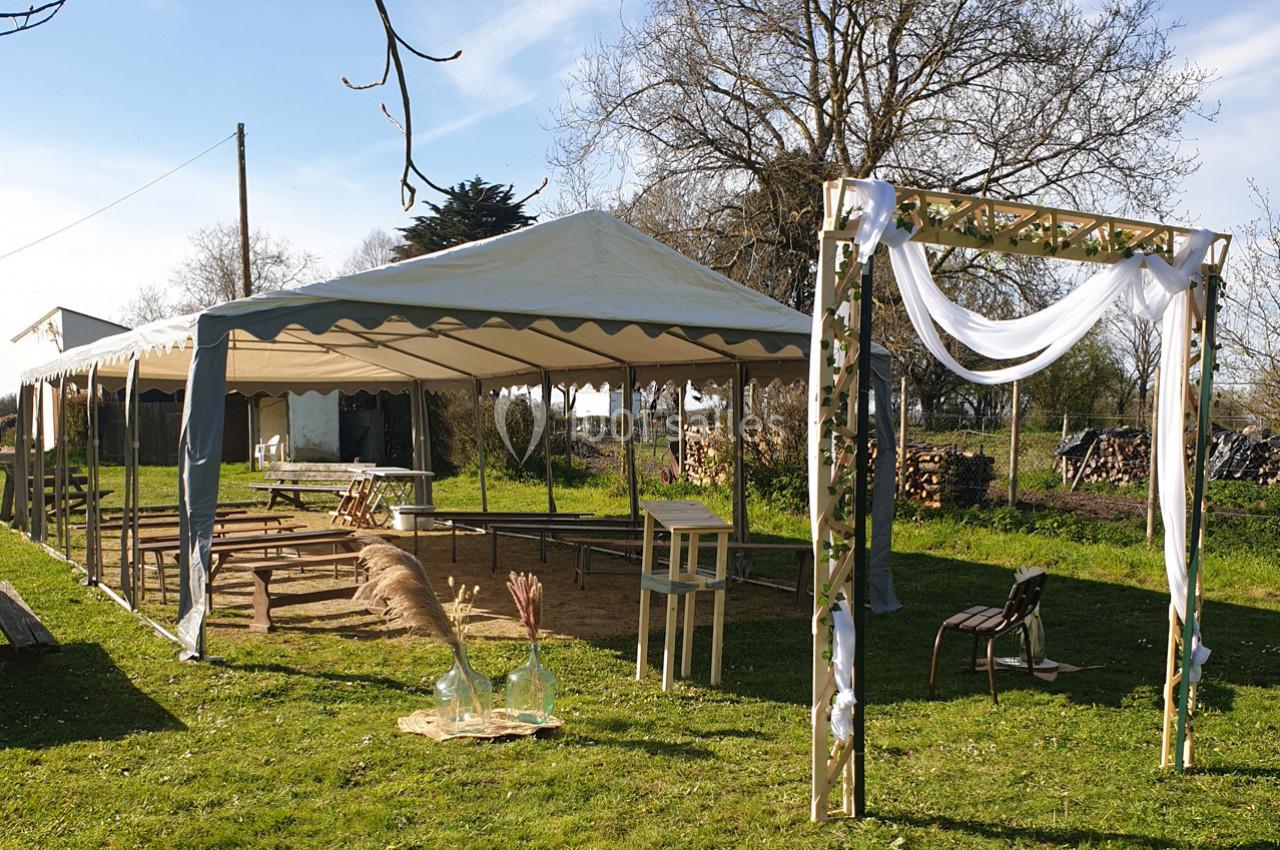 Tente blanche montée dans un jardin avec arche décorée et bancs en bois pour un événement en extérieur.