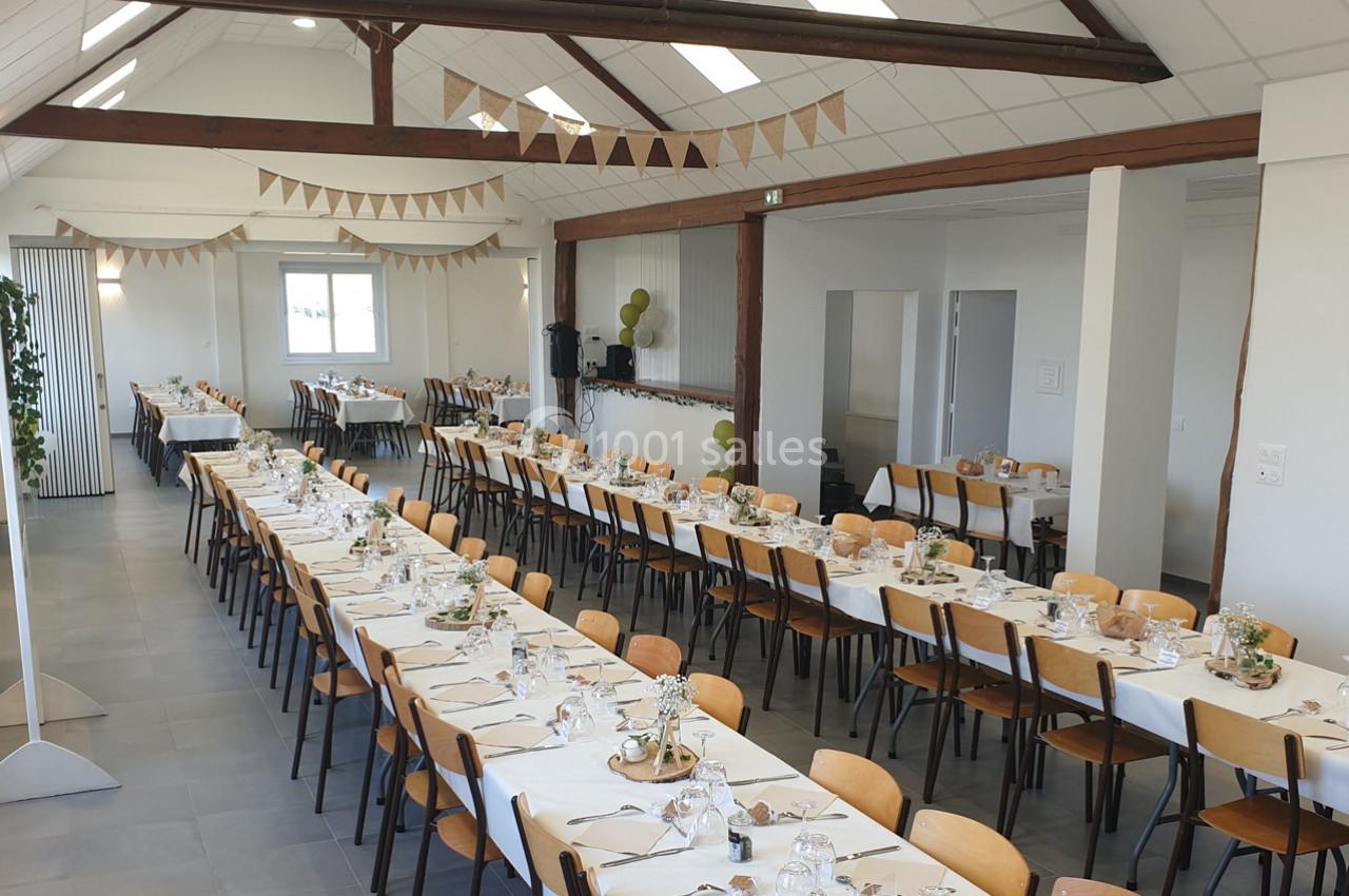 Salle de réception décorée avec des tables alignées, nappes blanches, chaises en bois et guirlandes suspendues.