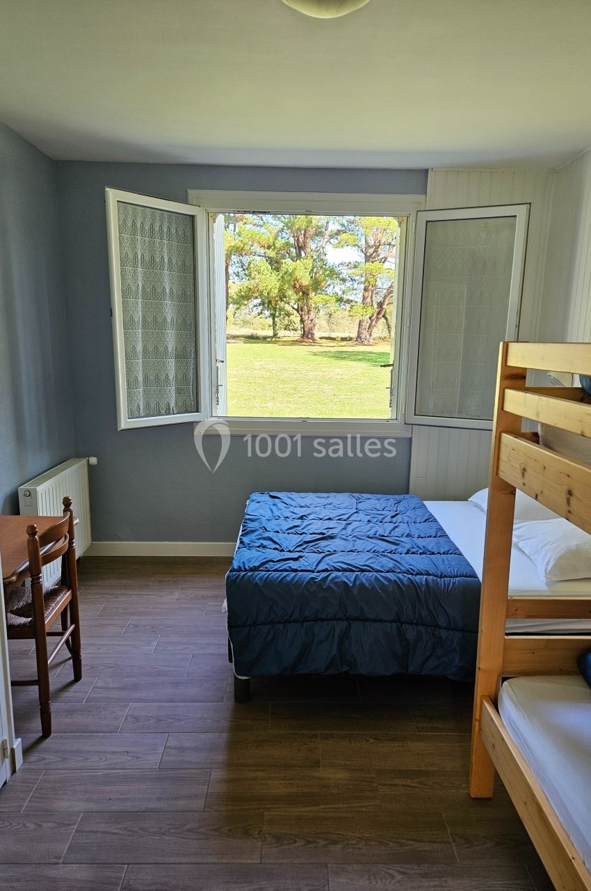 Chambre lumineuse avec lit double, lits superposés, chaise en bois et fenêtre ouverte donnant sur un jardin verdoyant.