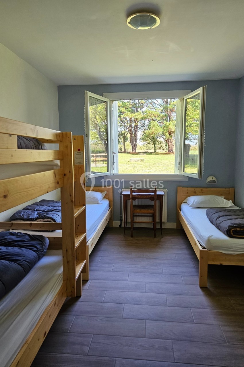 Chambre simple avec lits superposés, lit simple, bureau et fenêtre ouverte donnant sur un paysage arboré.