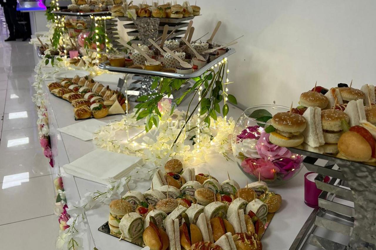 Buffet de mini-sandwichs variés et desserts disposés sur une table décorée de fleurs et de lumières.