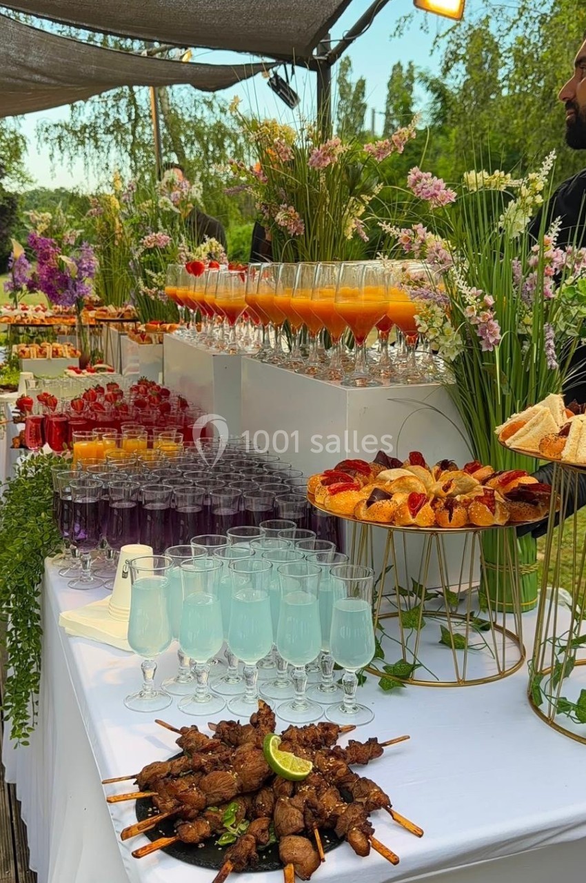 Buffet en extérieur avec brochettes, cocktails colorés, viennoiseries et décor floral sur une table blanche.