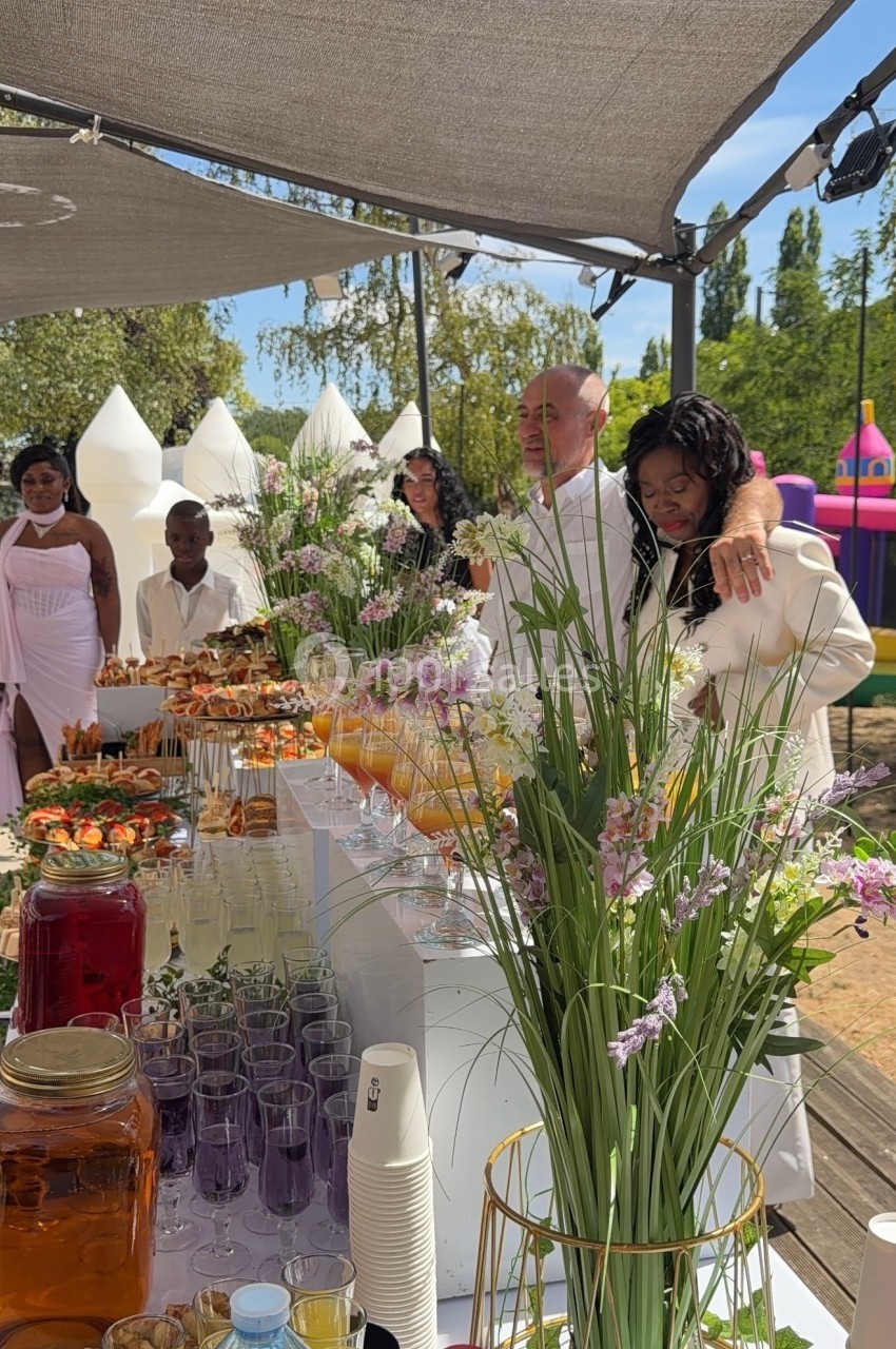 Table garnie de boissons et de plats colorés lors d'une réception en plein air, avec des invités en arrière-plan.