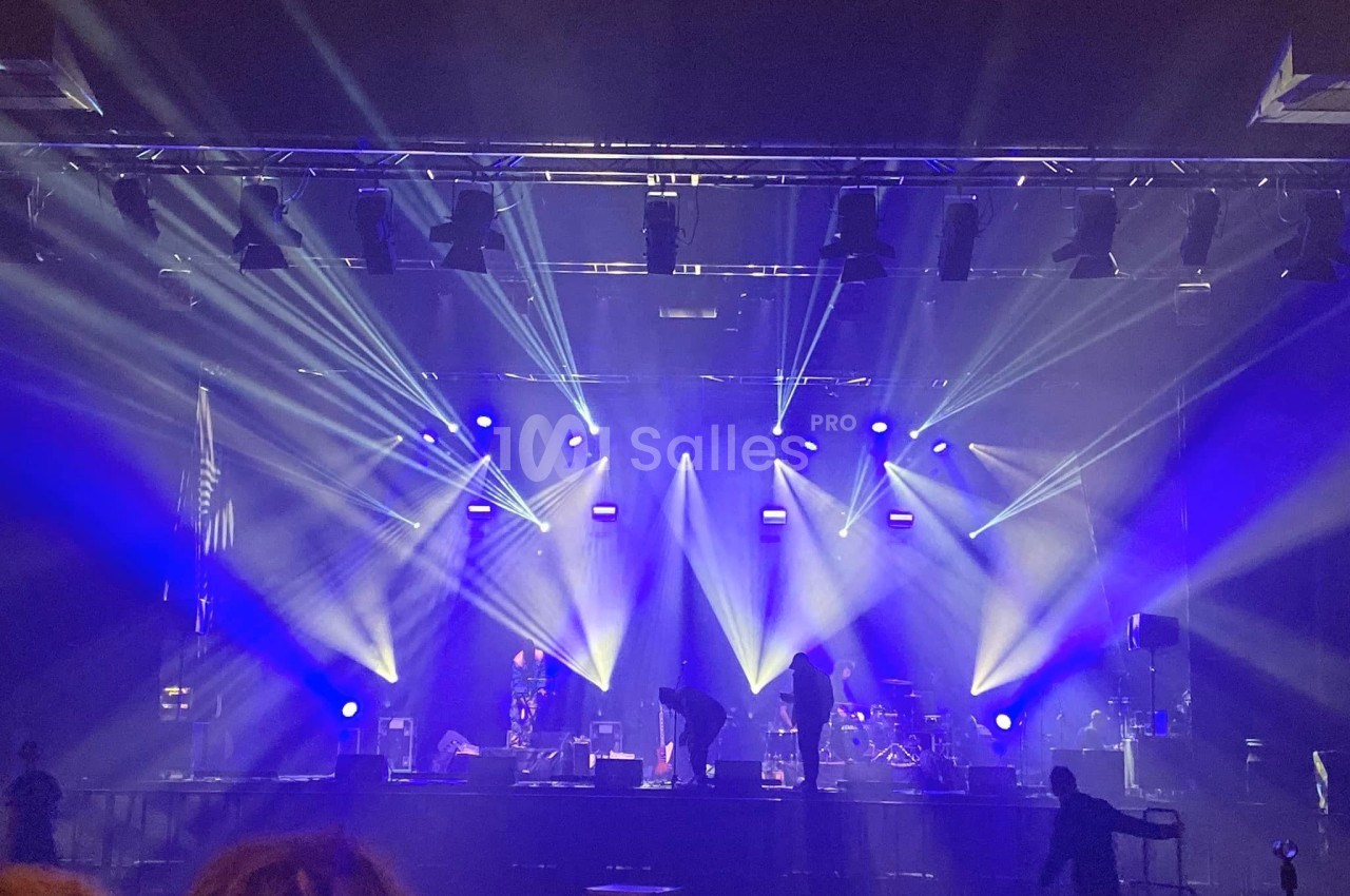 Scène de concert en intérieur avec éclairage bleu et techniciens préparant l'installation avant un spectacle.