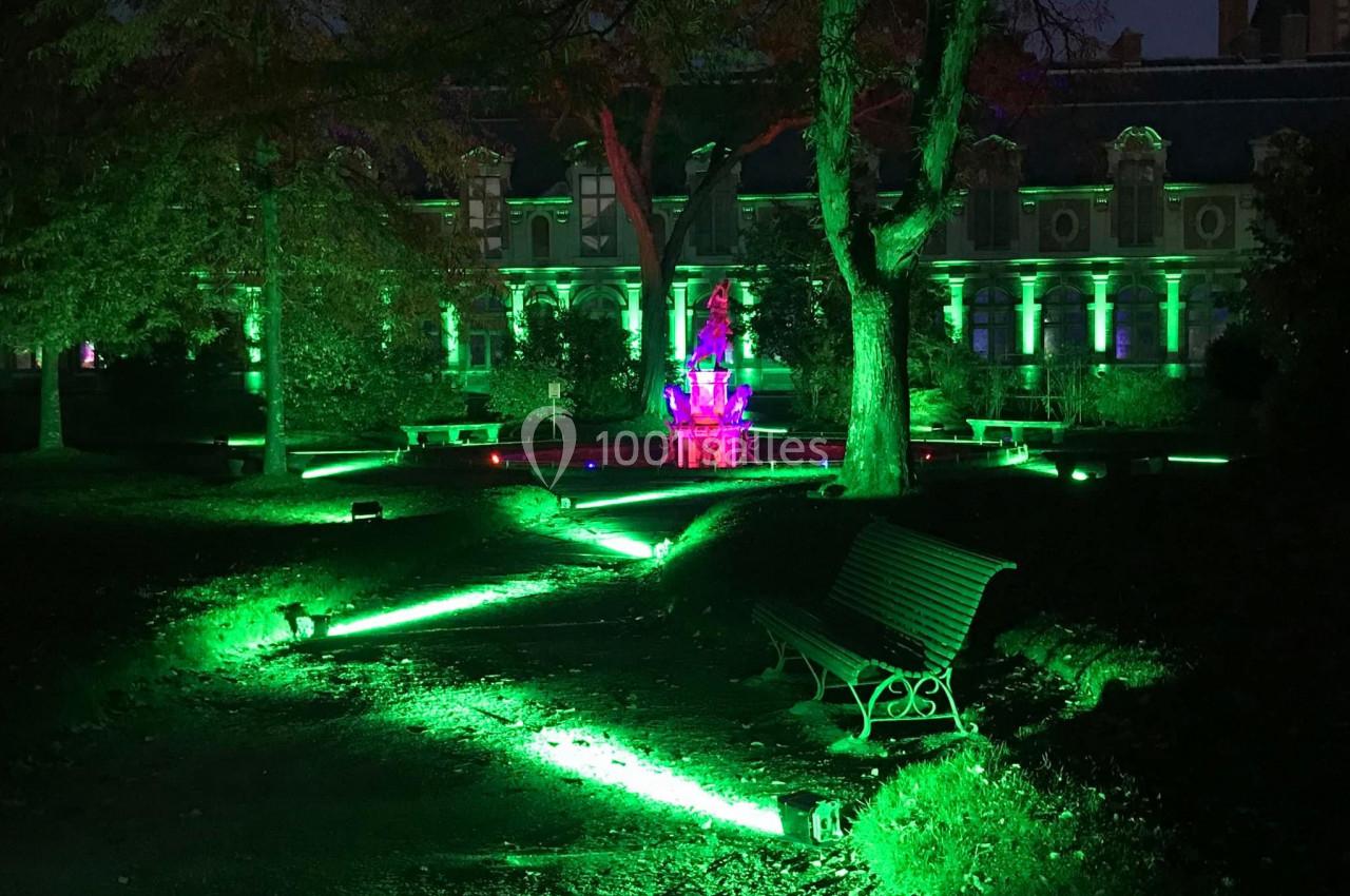 Éclairage nocturne vert illuminant un jardin avec une fontaine centrale et un bâtiment en arrière-plan.