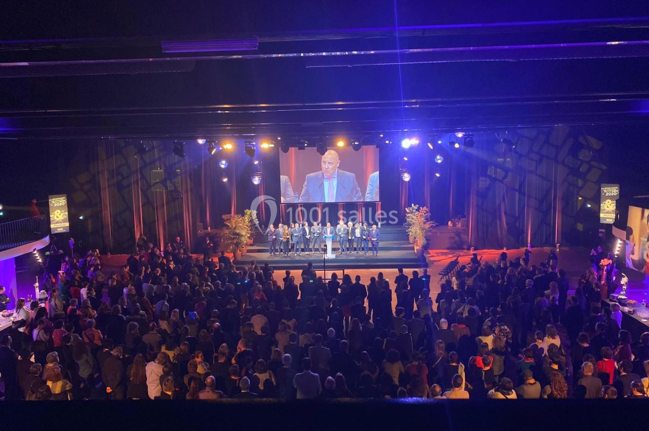 Vue d'une cérémonie sur scène avec un orateur, des participants alignés et une audience nombreuse dans une salle éclairée.