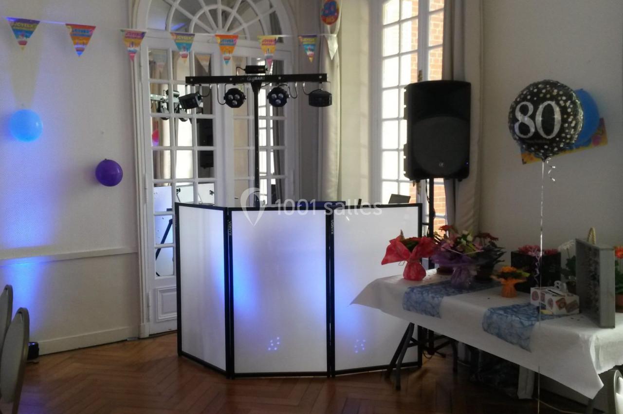 Salle décorée pour une fête avec un stand de DJ éclairé, des ballons, des guirlandes et une table ornée de fleurs.