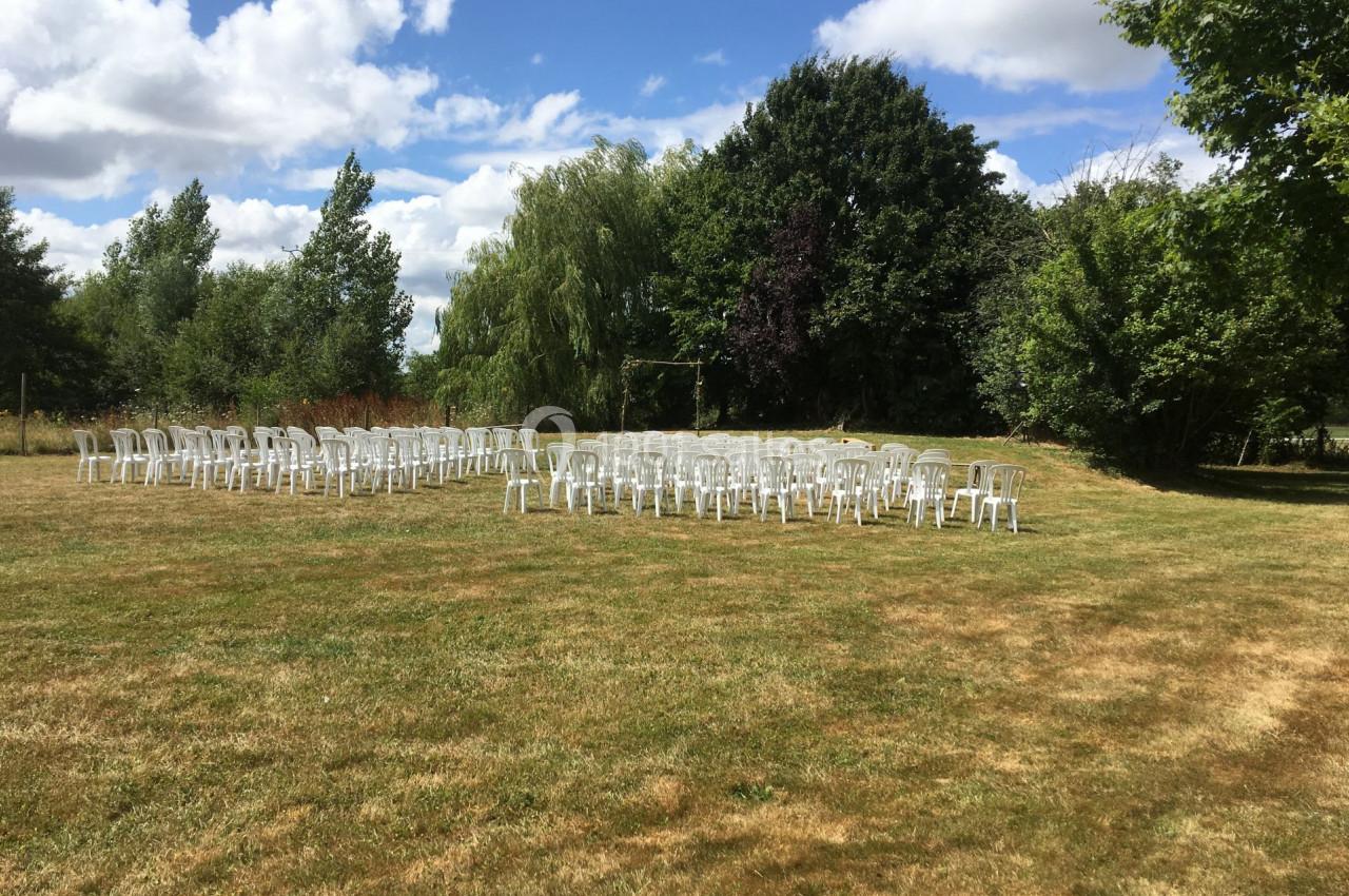 Chaises blanches disposées en rangées sur une pelouse, entourées d'arbres sous un ciel partiellement nuageux.