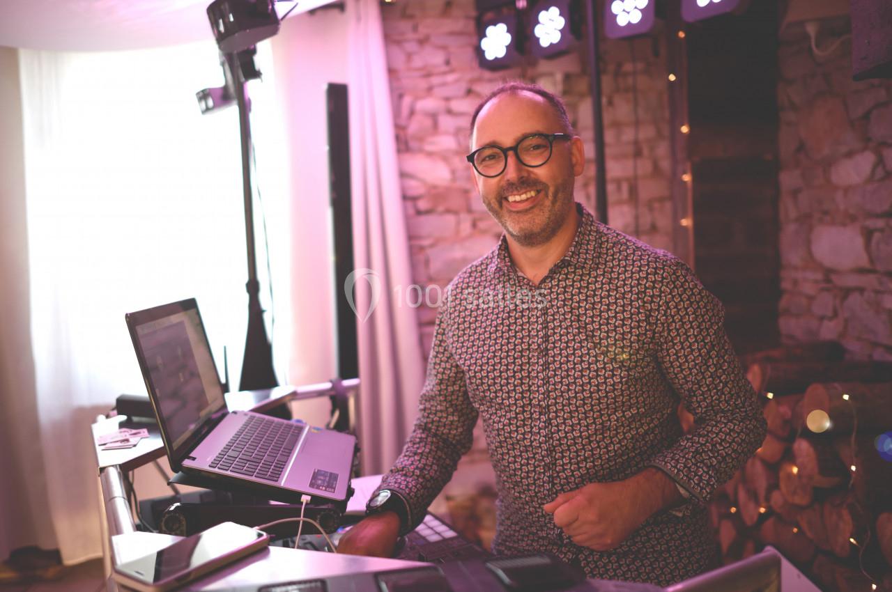 Un homme souriant se tient derrière une table avec un ordinateur portable, dans un environnement lumineux et chaleureux.