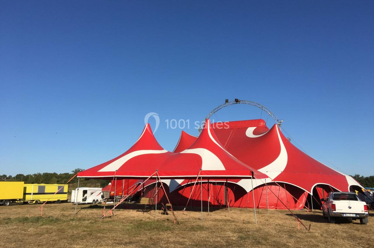Grand chapiteau rouge et blanc monté sur un terrain herbeux, entouré de véhicules et d'un ciel dégagé.