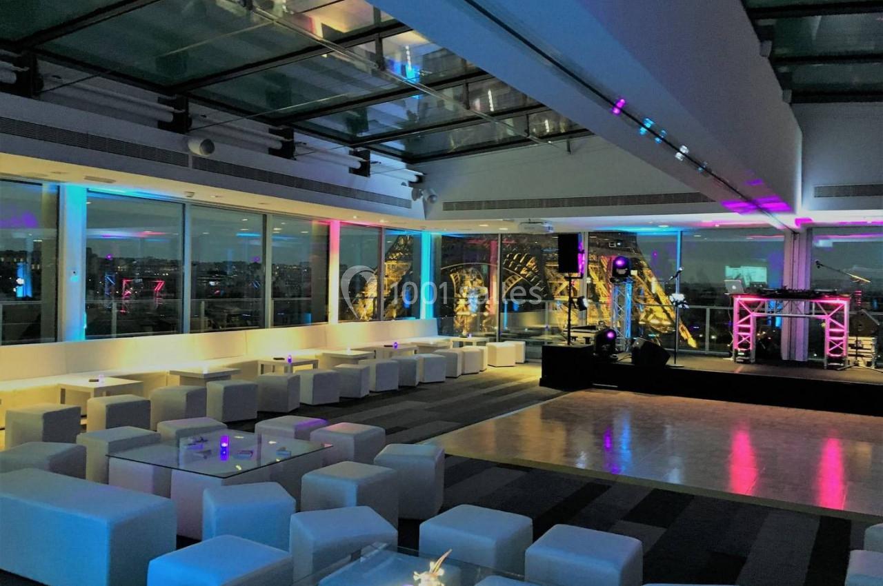 Salle moderne avec mobilier blanc, éclairage coloré et vue sur la tour Eiffel à travers de grandes baies vitrées.