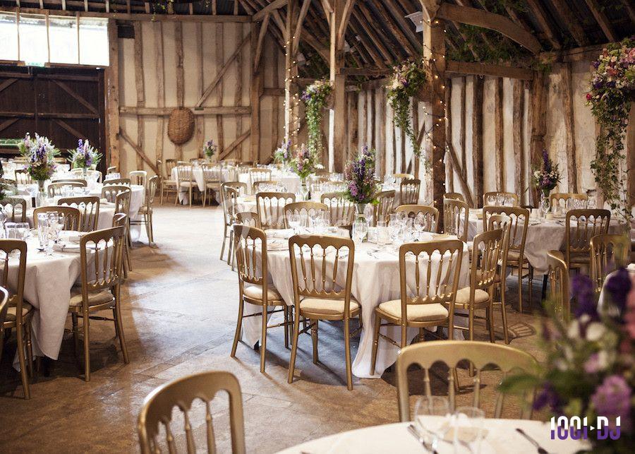 Salle de réception rustique avec tables rondes dressées, chaises en bois et décorations florales.