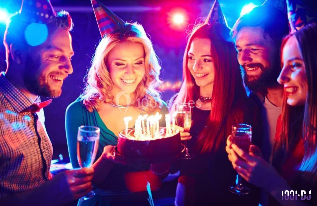 Un groupe de personnes souriantes fête un anniversaire avec un gâteau illuminé par des bougies et des coupes de champagne.