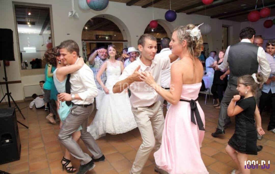 Des invités dansent lors d'une fête, avec une mariée en arrière-plan dans une salle décorée de lanternes colorées.