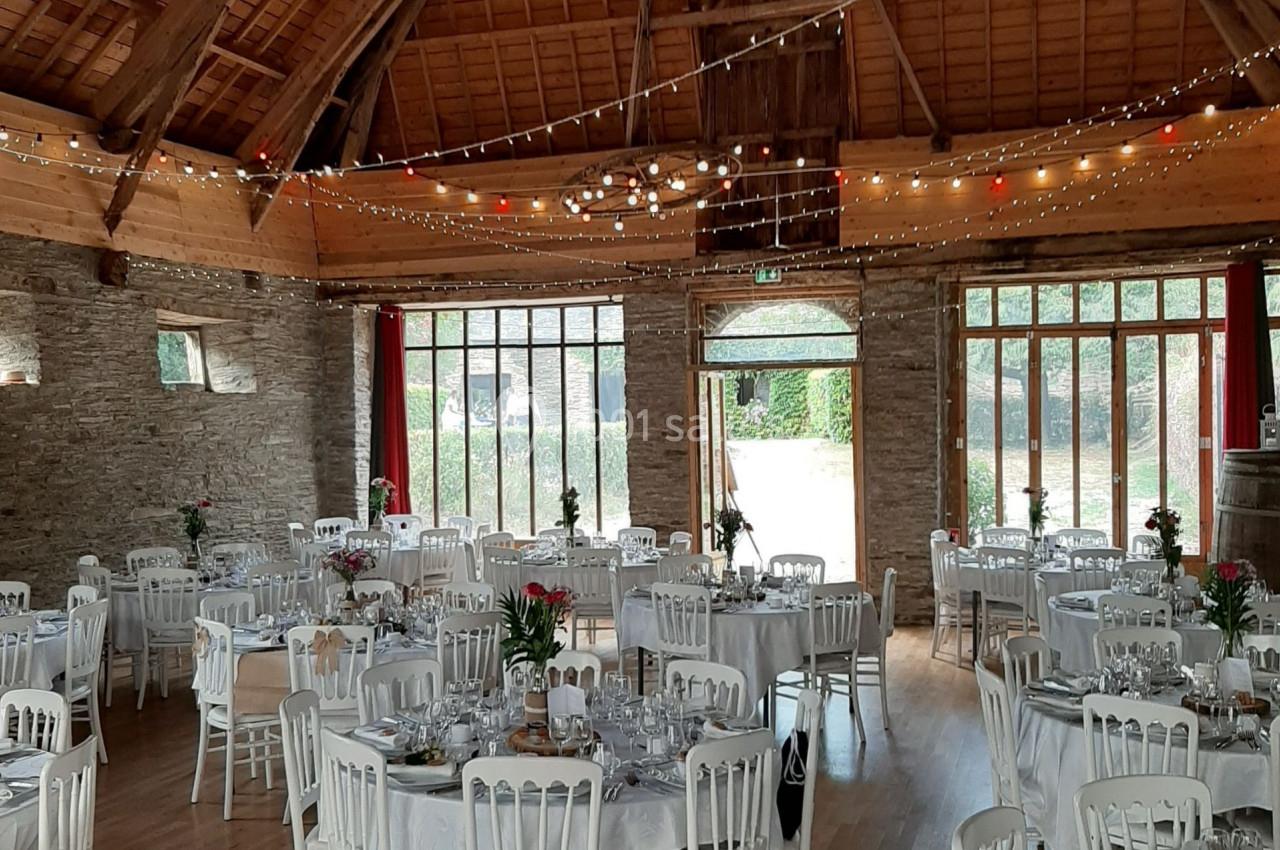 Salle de réception en pierre avec tables rondes dressées, chaises blanches et guirlandes lumineuses suspendues.