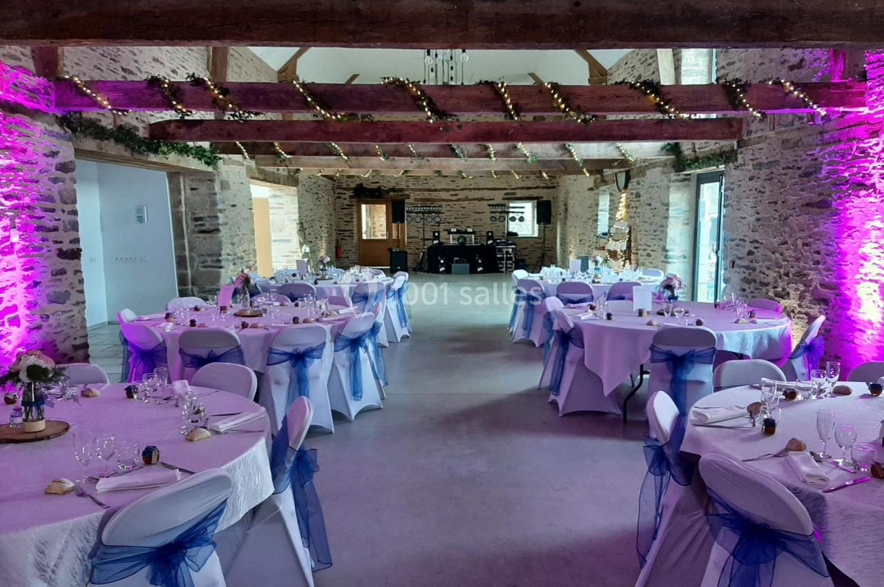 Salle de réception décorée avec des tables rondes, nappes blanches, chaises ornées de rubans bleus et éclairage rose.