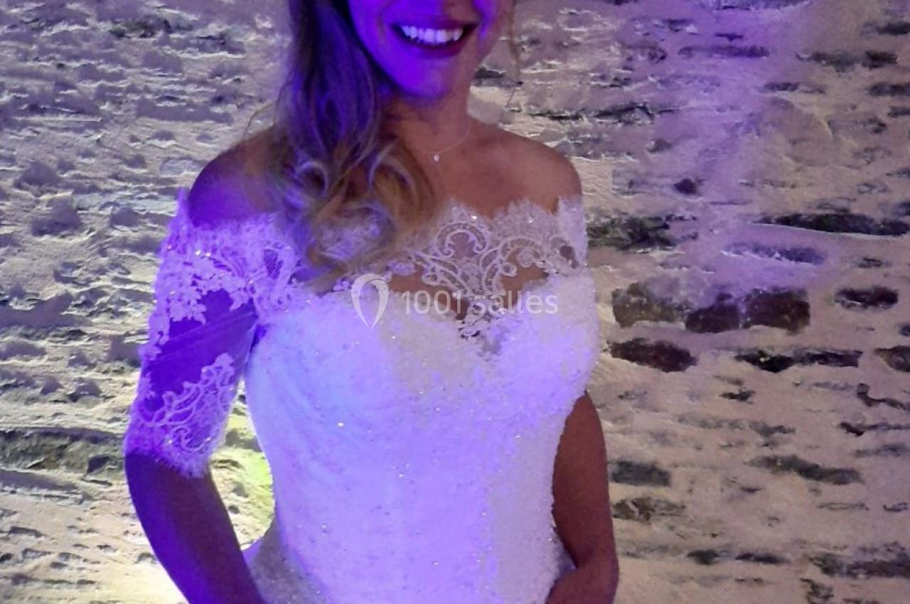 Femme souriante en robe de mariée blanche avec dentelle, posant devant un mur en pierre éclairé par une lumière violette.