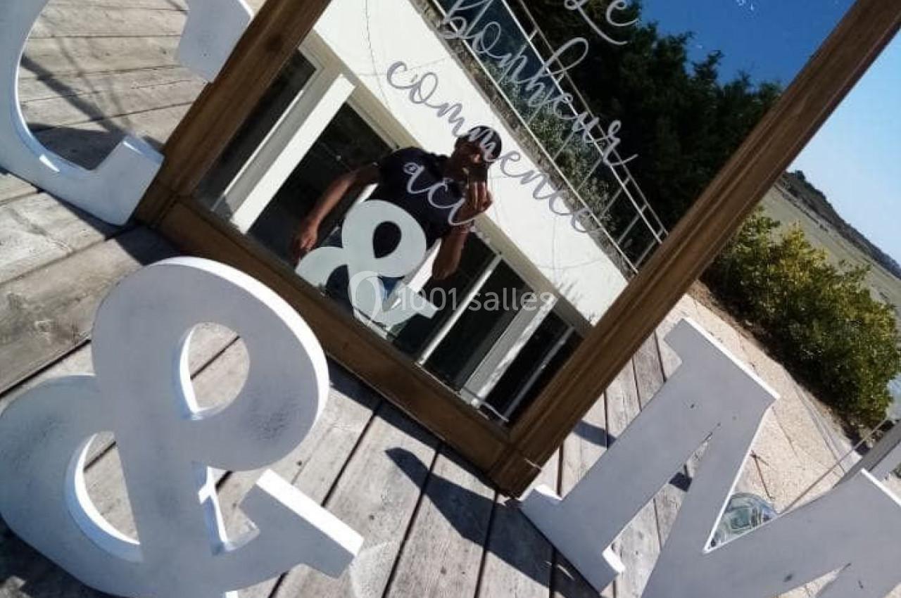 Un miroir avec une inscription réfléchit un photographe, entouré de grandes lettres décoratives sur une terrasse en bois.