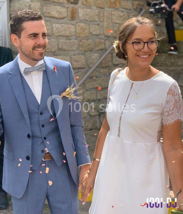 Un couple souriant en tenue de mariage, tenant la main, entouré de pétales de fleurs devant un mur en pierre.