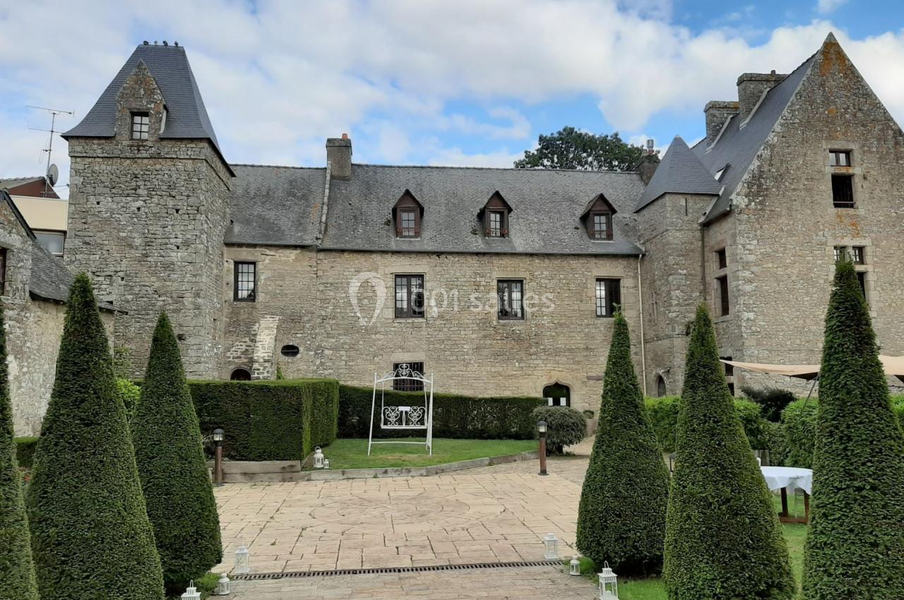 Façade en pierre d'un manoir historique entouré de conifères taillés et d'une cour pavée.