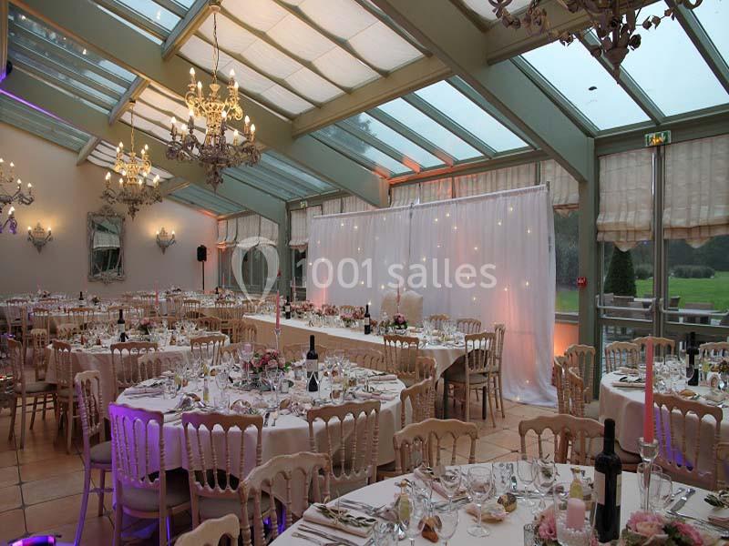 Salle de réception lumineuse avec tables décorées, chaises en bois clair, lustres et grandes baies vitrées donnant sur un…
