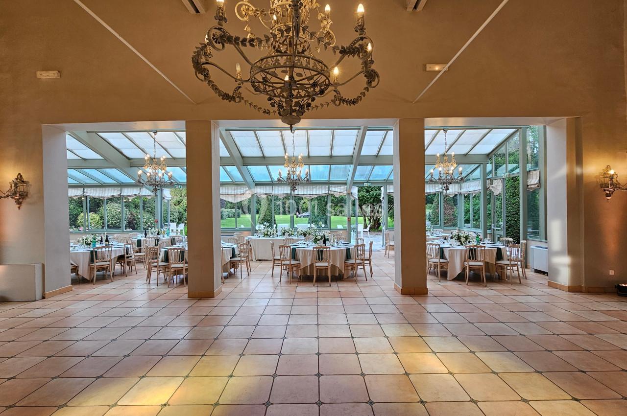 Salle de réception lumineuse avec grandes baies vitrées, vue sur un jardin, tables dressées pour un événement.