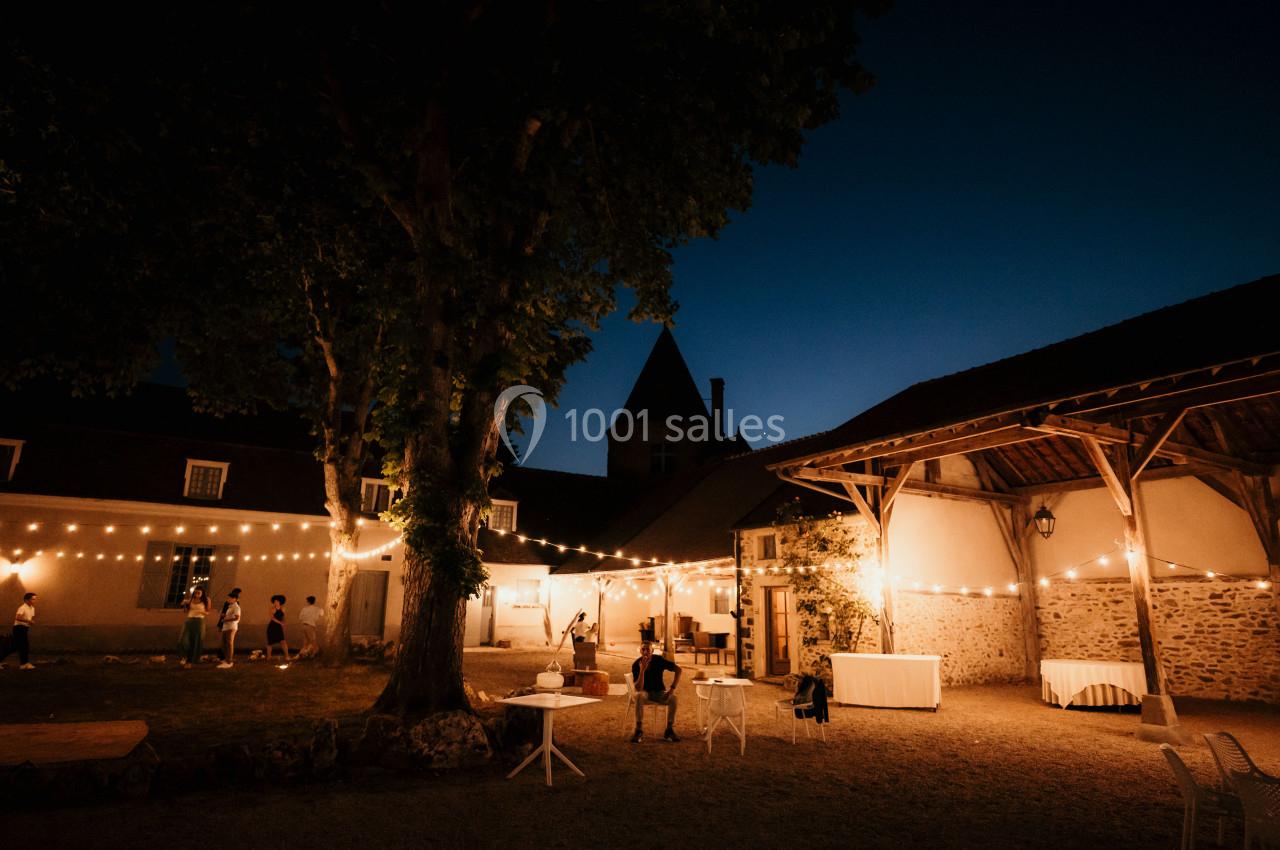 Cour extérieure éclairée par des guirlandes lumineuses au crépuscule, avec quelques personnes et bâtiments en arrière-plan.