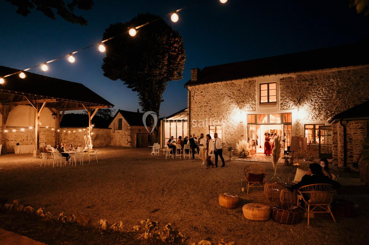 Cour d'une bâtisse en pierre éclairée la nuit, avec des invités assis ou debout sous des guirlandes lumineuses.