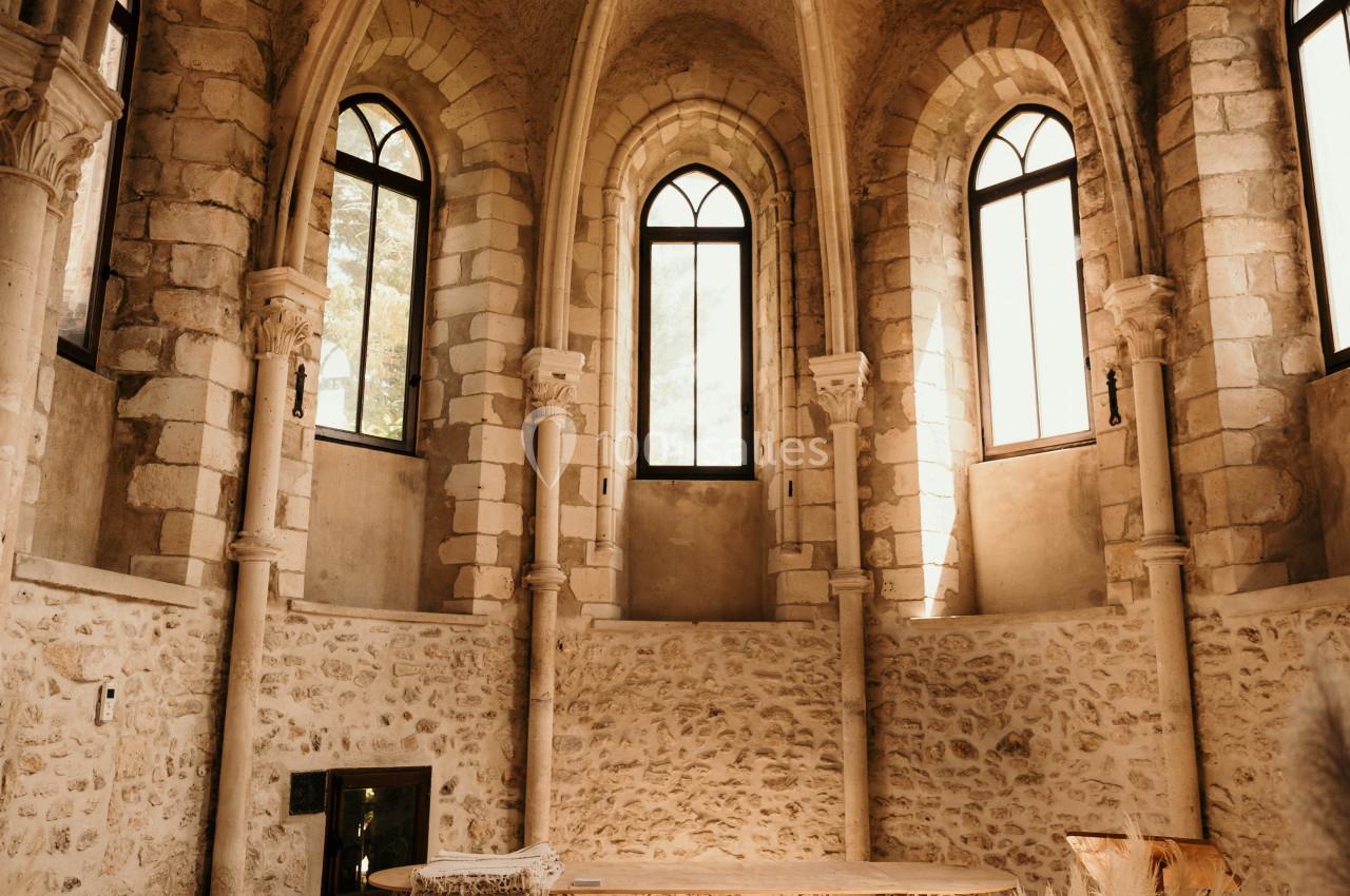 Salle voûtée en pierre avec grandes fenêtres cintrées, mobilier en fer forgé et décoration florale lumineuse.