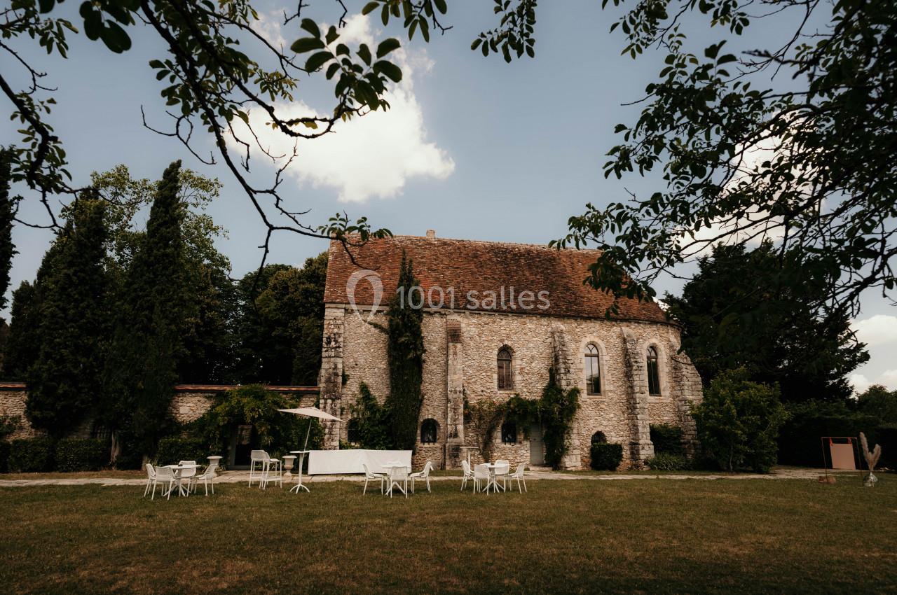 Vue d'une ancienne bâtisse en pierre entourée d'arbres, avec des tables et chaises blanches disposées sur une pelouse.