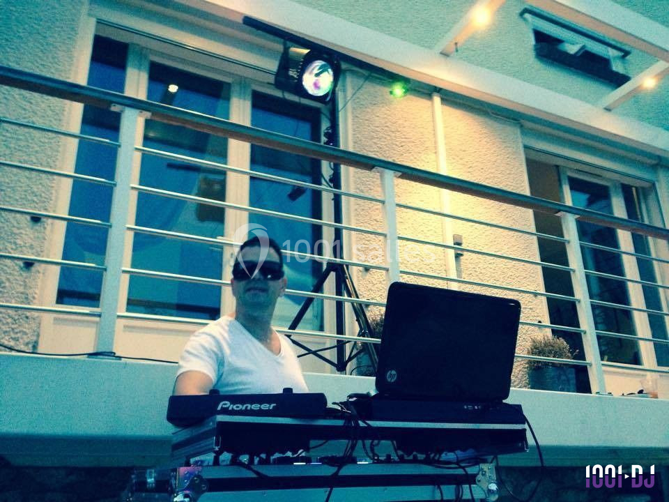 Un DJ en extérieur devant une maison, équipé de matériel de mixage et d'un ordinateur portable.