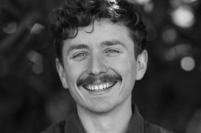 Portrait en noir et blanc d'un homme souriant avec des cheveux bouclés et une moustache, sur un fond flou naturel.