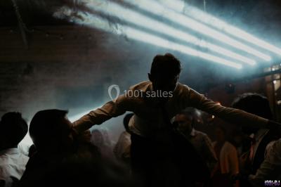 Homme souriant avec une moustache, portant une chemise sombre, devant un arrière-plan flou de verdure.
