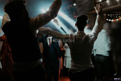Homme souriant avec une moustache, portant une chemise sombre, devant un arrière-plan flou de verdure.